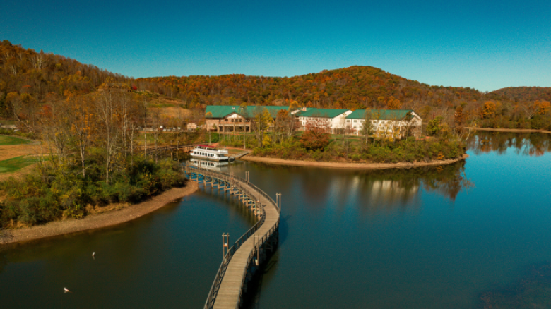 A scenic view of a lake with a curved boardwalk leading to a building surrounded by autumn trees and hills under a clear blue sky.