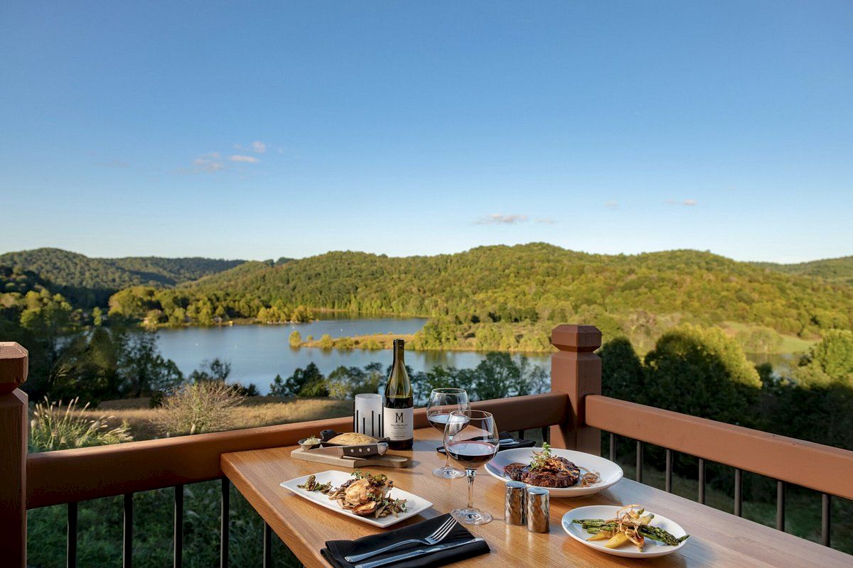 A scenic view of a lake and forested hills with a table set for dining, featuring wine, glasses, and plates of food on a wooden deck.