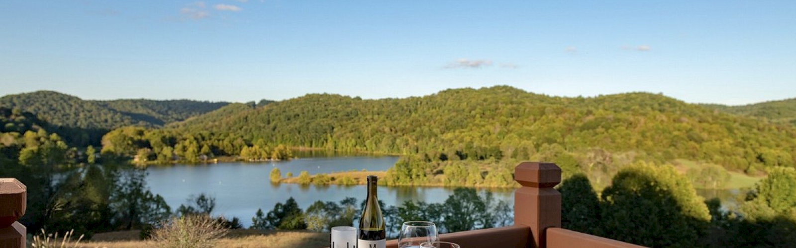 A scenic outdoor dining setup with wine, two plates of food on a wooden table, overlooking a lake and lush green hills under a clear blue sky.